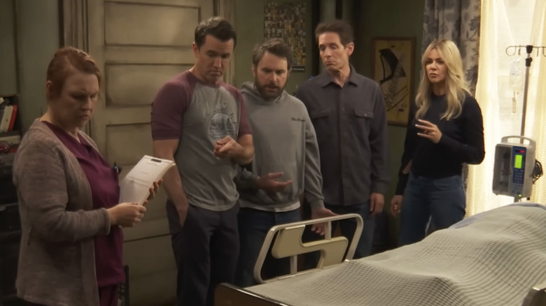 Mac, Charlie, Dennis, and Dee stand at someone's bedside in Charlie's apartment on It's Always Sunny in Philadelphia