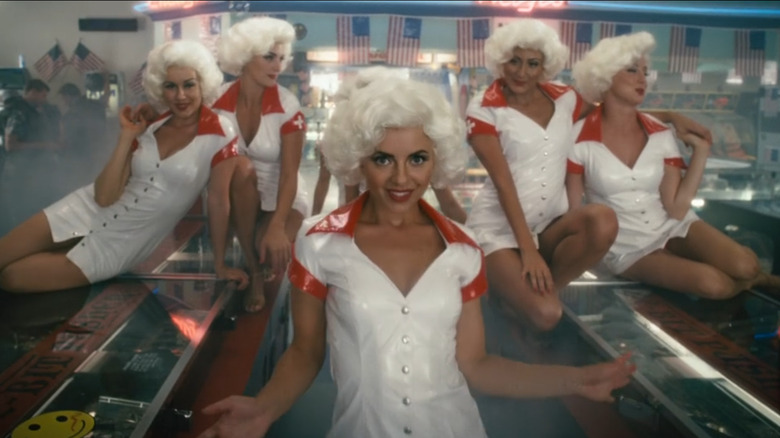 A group of dancers in Marilyn Monroe-esque wigs and latex nurses outfits pose on arcade machines in Southland Tales