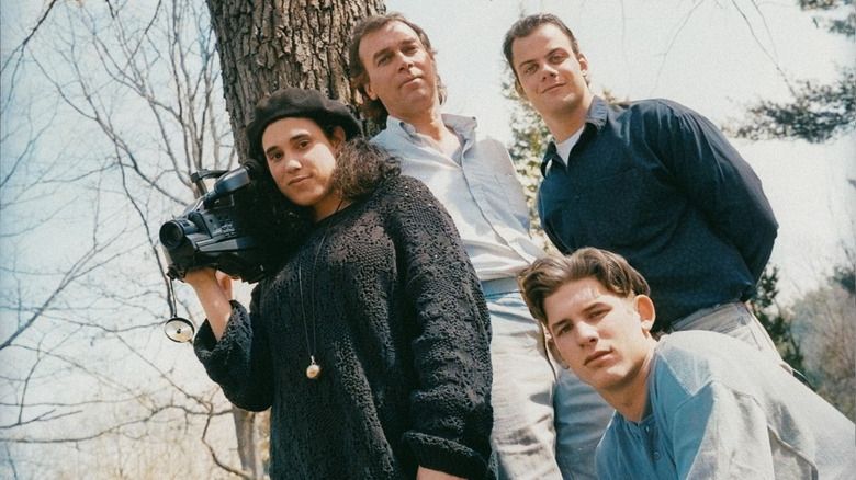 Three high school students, one carrying a camcorder, pose for a photo with their teacher outside in Teenage Wasteland