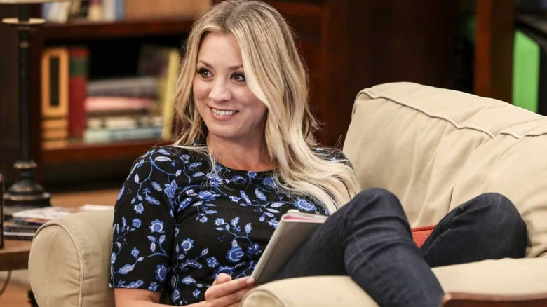 Penny is sitting on the couch, grinning, in The Big Bang Theory