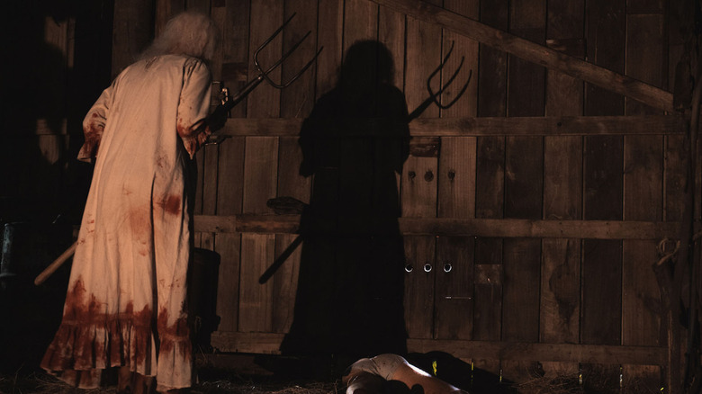 woman in a bloody night dress holding a pitchfork in a dark barn next to a man on the floor in his underwear