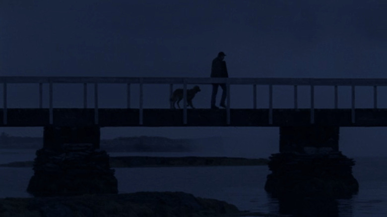 Tom Selleck's Jesse Stone walks across a walkway over some water at dusk in Jesse Stone Stone Cold