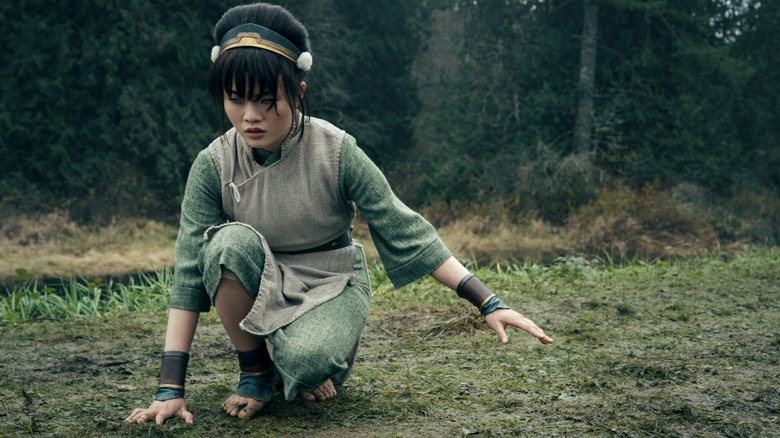 Miya Cech as Toph kneeling on the ground outside of a forest in Avatar: The Last Airbender