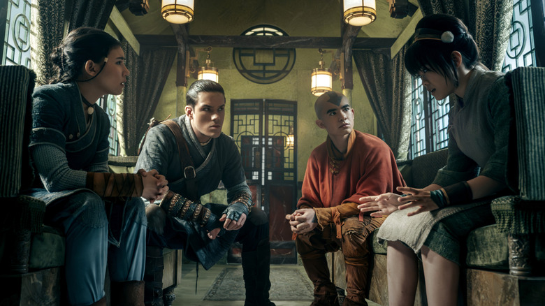 Kiawentiio Tarbell as Katara, Ian Ousley as Sokka, Gordan Cormier as Aang, and Miya Cech as Toph sitting together in an ornate room in Avatar: The Last Airbender