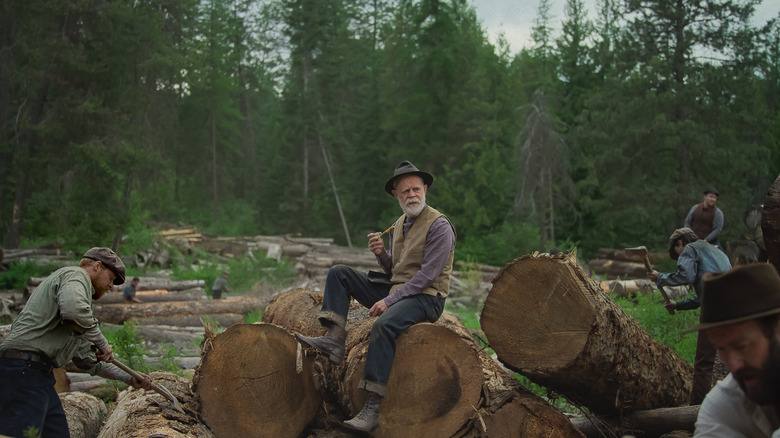 Arn sits on a log and smokes a pipe while others work around him in Train Dreams