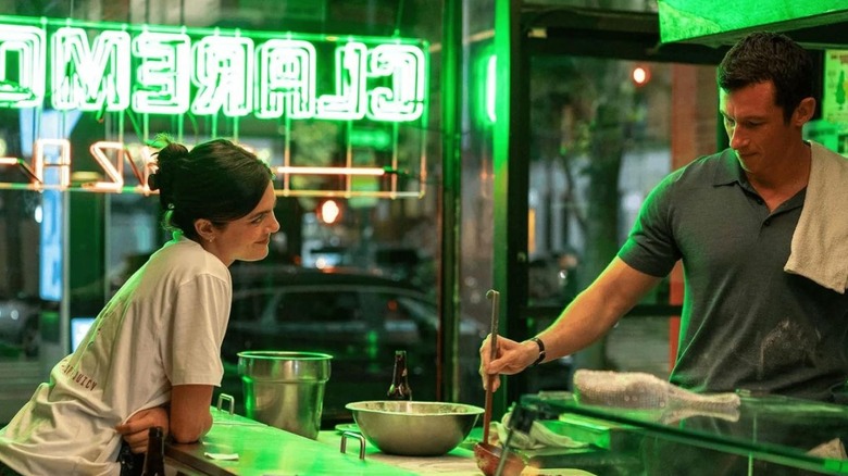 Monica Barbaro as Allie standing at the counter talking to Callum Turner as Owen as he makes a pizza in One Night Only