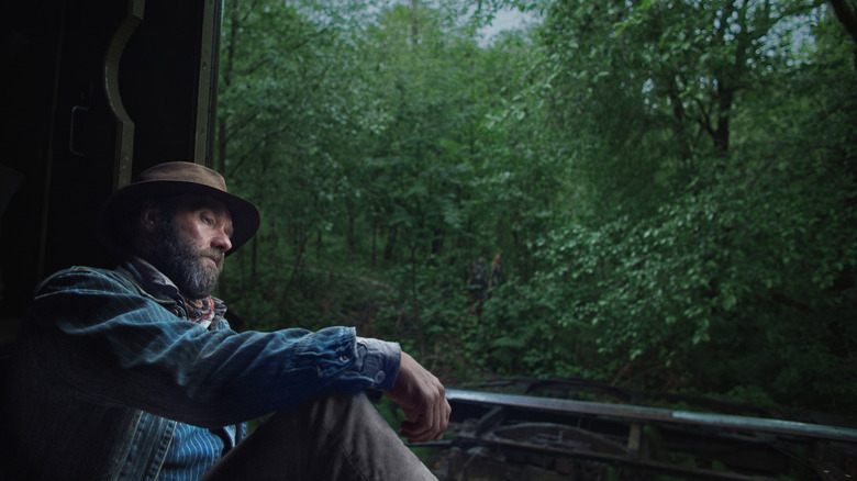 Joel Edgerton as Robert Grainier in Train Dreams sitting in a train car with a forest outside