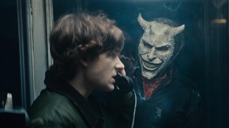 Ethan Hawke as The Grabber, looking on at Mason Thames as Finney Blake talking on a black phone in a phone booth in Black Phone 2.