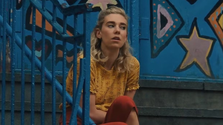 Alina (Vanessa Kirby), in a yellow patterned shirt and red pants, sits distraught on a city staircase, with blue doors covered in brightly colored shapes behind her in Italian Studies