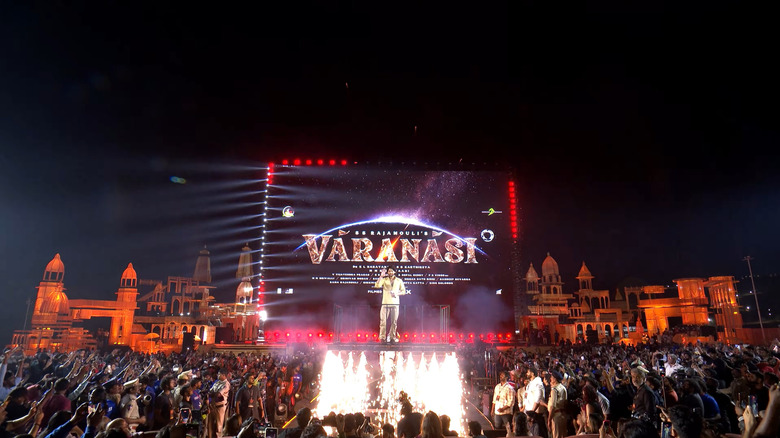 Actor Mahesh Babu standing on top of a platform in front of a crowd during the announcement event for Varanasi