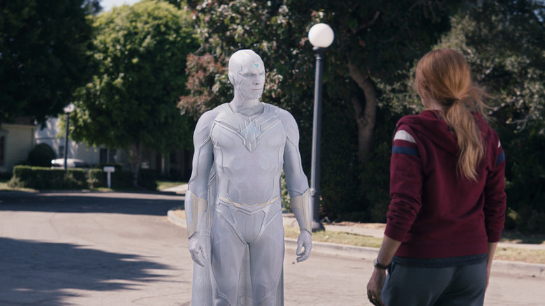 Paul Bettany as White Vision standing on a suburban street next to Elizabeth Olsen as Wanda Maximoff in WandaVision