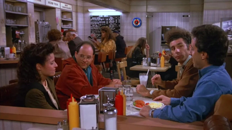 Julia Louis-Dreyfus as Elaine, Jason Alexander as George, Michael Richards as Kramer, and Jerry Seinfeld as Jerry sitting at a diner table on Seinfeld