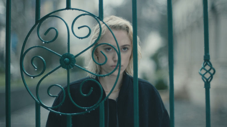 blonde woman standing behind a green iron fence in a black jacket