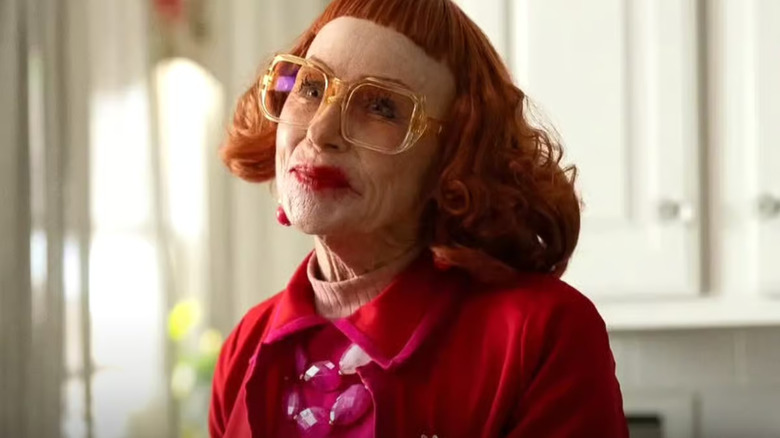 Gladys in yellow glasses wearing her wig and smirking in Marcus' kitchen in Weapons