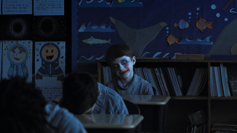 Alex Lilly with creepy makeup sits in a dark classroom while other students have their heads on their desks in Weapons