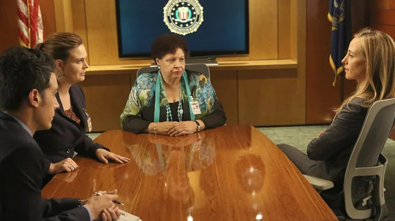 Caroline Julianne, played by Patricia Belcher, sits at the head of the table alongside Temperance Brennan, played by Emily Deschanel, and other attendees in a government office in Bones.