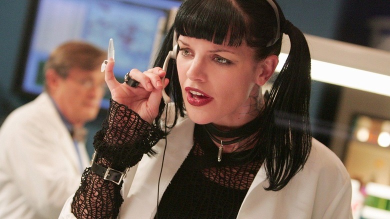 Abby, wearing a vial on her pinky and holding a small, high-tech phone device, wearing a lab coat and porting her usual Goth garb. A scene from NCIS.