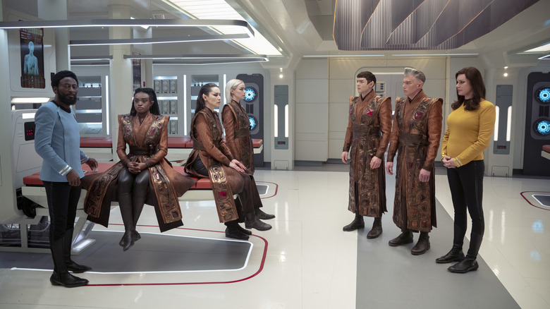 Babs Olusanmokun as Doctor M'Benga and Rebecca Romijn as Una standing in the sick bay with Celia Rose Gooding as Uhura, Christina Chong as La'an, Jess Bush as Chapel, Ethan Peck as Spock, and Anson Mount as Pike dressed in Vulcan attire in Star Trek: Strange New Worlds