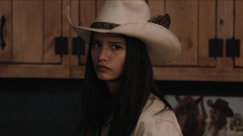 Avery stands in the Yellowstone bunkhouse wearing a white cowboy hat looking confrontational