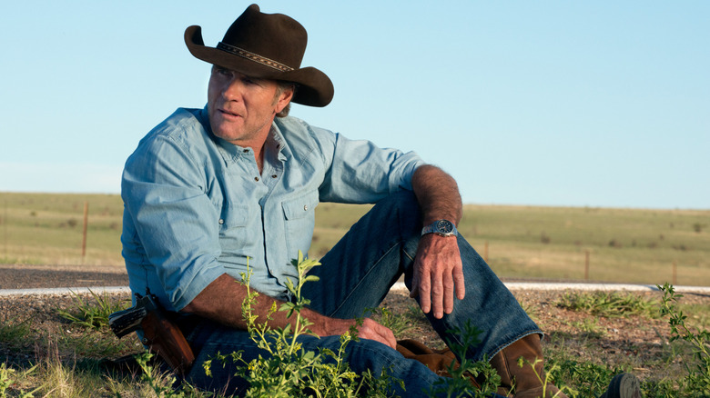 Sheriff Walt Longmire (Robert Taylor) sits on the grass by a road in Longmire
