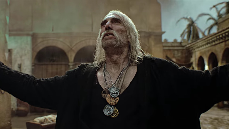 Sharlto Copley as Leo Bonhart spreading his arms while wearing many Witcher pendants on The Witcher