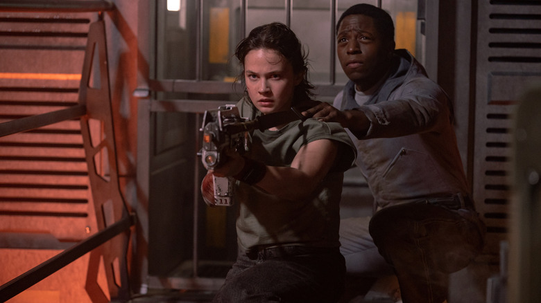Cailee Spaeny as Rain holding a futuristic gun while David Jonsson as Andy crouches protectively behind her in Alien: Romulus