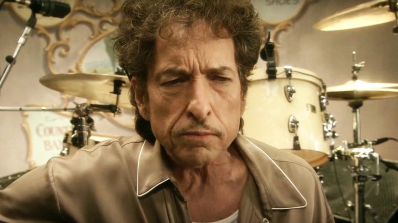 Bob Dylan as Jack Fate sits in a chair with his guitar as he sings a song in Masked and Anonymous