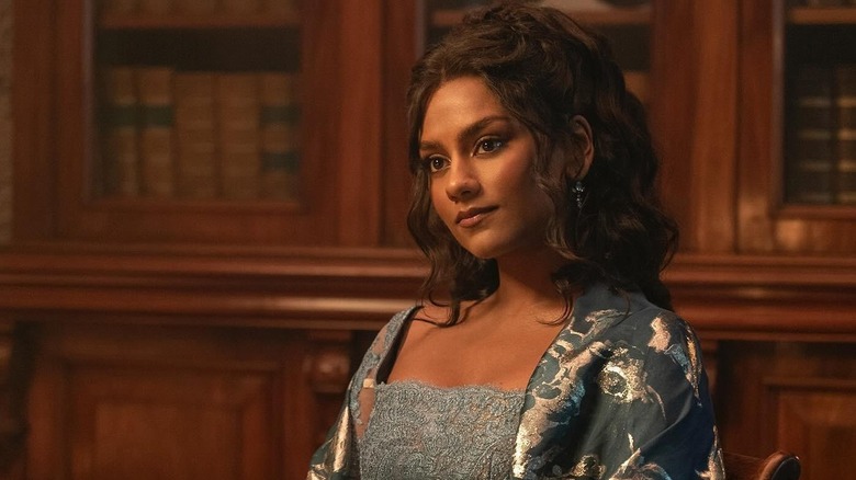 Simone Ashley as Kate Sharma sitting in a chair in front of a bookcase in Bridgerton