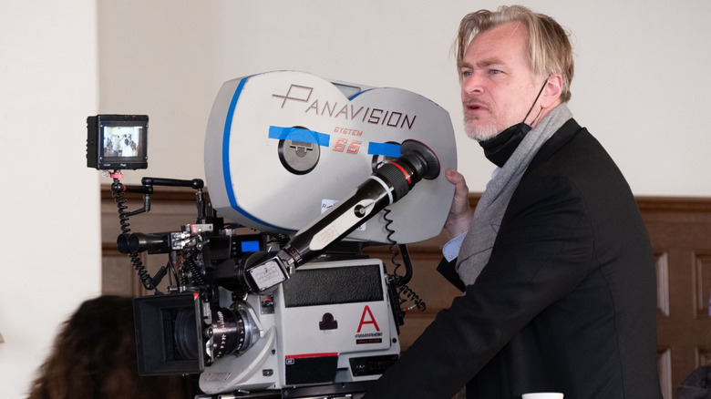 Christopher Nolan stands behind a camera with a face mask around his neck on the Oppenheimer set