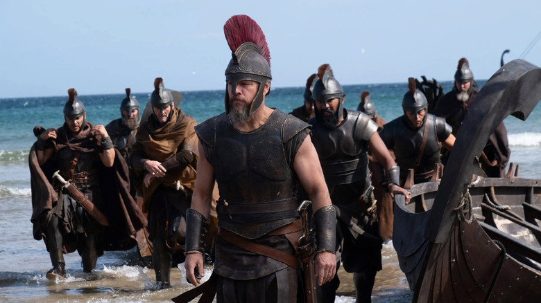 Matt Damon as Odysseus wearing a helmet and walking on a beach near a boat in front of Himesh Patel as Eurylochus and several other soldiers in The Odyssey