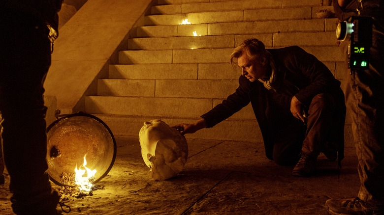 Director Christopher Nolan carefully adjusting the head of a fallen statue in front of a staircase on the set of The Odyssey