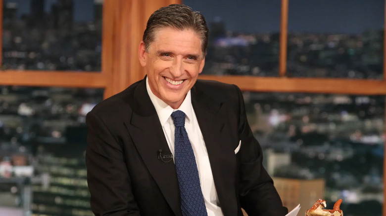 Craig Ferguson smiles cheerfully wearing a suit and tie behind the desk of The Late Late Show