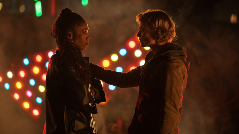 Tom Rhys Harries as Matt Hagen holding his arm out and touching the shoulder of Naomi Ackie as Caitlyn Corr in front of a colorful sign in Clayface