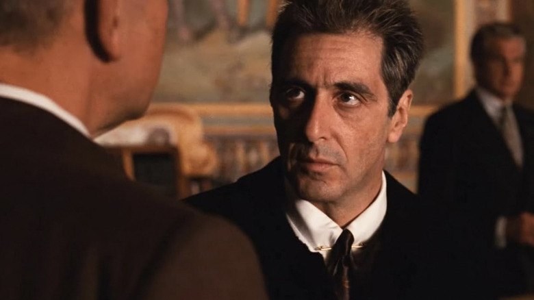 தி காட்பாதர் பகுதி III இல் வின்சென்ட் கார்லியோன் பின்னணியில் கீழே பார்க்கும் ஆண்டி கார்சியாவாக மைக்கேல் கோர்லியோனாக சாம்பல் நிறத்தில் சன்கிளாஸ் அணிந்த அல் பசினோ