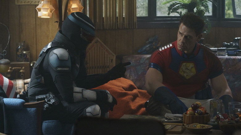 Freddie Stroma as Vigilante in costume and John Cena as Peacemaker in costume sitting together in Peacemaker season 1