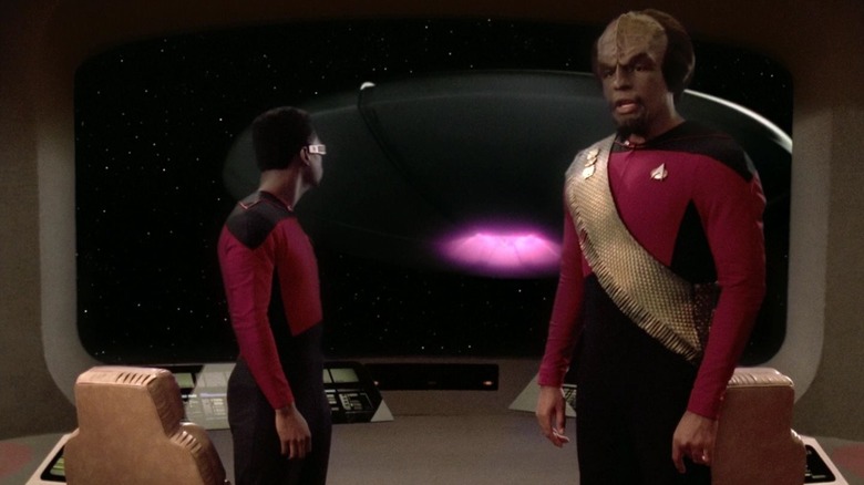 Worf and Geordi on the bridge of the U.S.S. Enterprise, Geordi is looking out of the viewscreen at a big, weird alien slying saucer. A scene from Star Trek: The Next Generation.