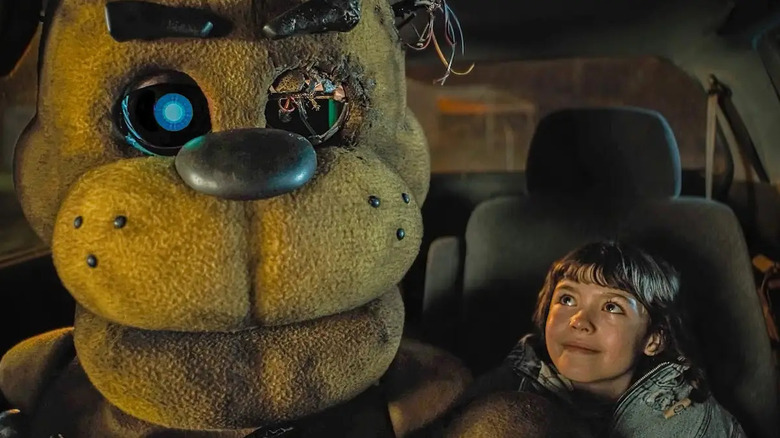 A broken Freddy Fazbear (Kevin Foster) sits and smiles in front of a smiling Abby (Piper Rubio) in the car in Five Night's at Freddy's (2023)