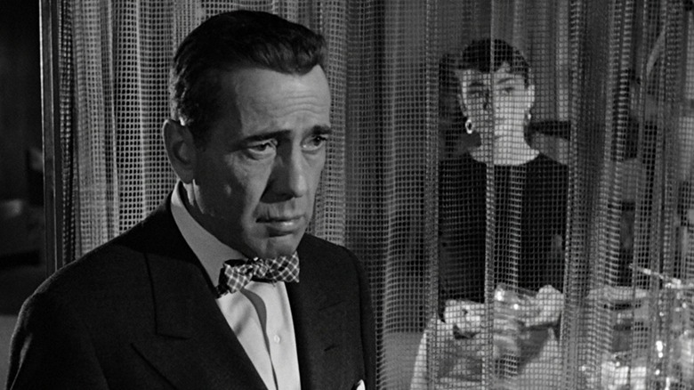 Humphrey Bogart's Linus Larrabee looks sad while Audrey Hepburn's Sabrina Fairchild is visible through a gauzy curtain in the background, in Sabrina