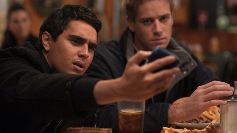 Divya Narendra holding up a phone confused while one of the Winklevoss twins looks concerned in The Social Network