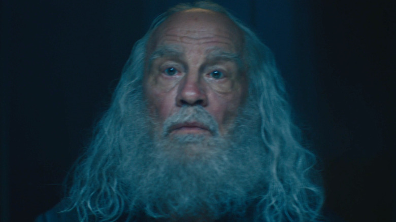 The Red Ghost stares mournfully past the camera with his long white hair and beard in a pitch black room in 