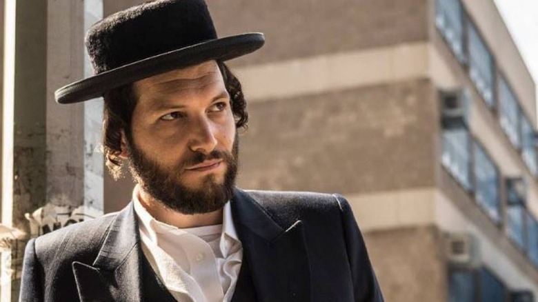 Noah wearing a hat on the street looking at something in Unorthodox