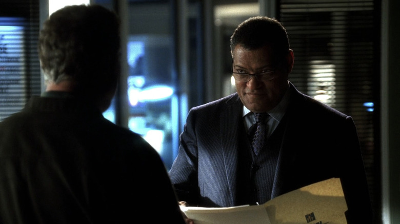 Laurence Fishburne mientras Raymond Langston sonríe mientras sostiene un archivo y se encuentra frente a alguien con la espalda a la cámara en CSI