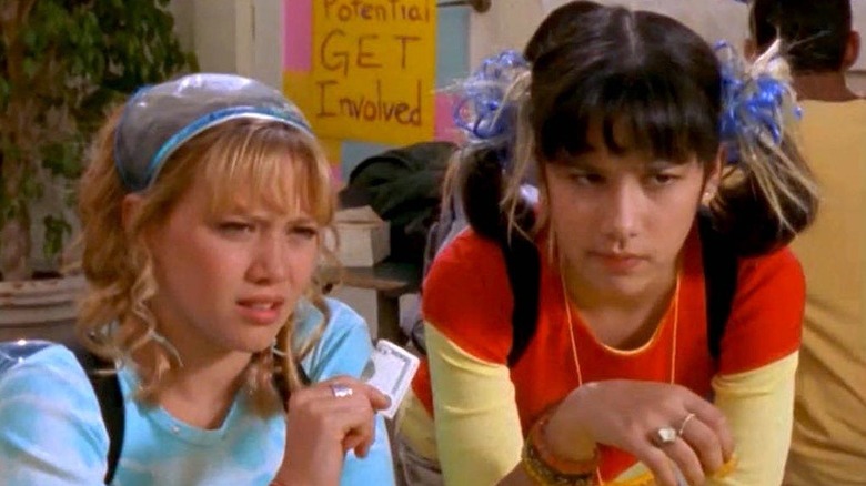 Lizzie holds folded money while she and Miranda, both wearing annoyed expressions, look to the right, dressed in colorful clothing with funky hair accessories in Lizzie McGuire