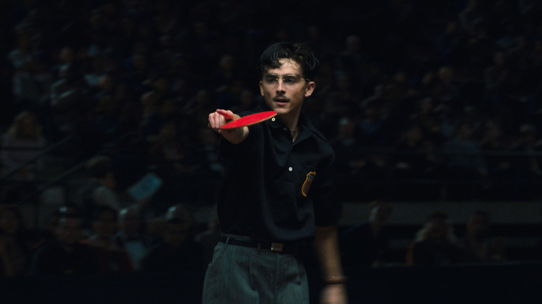 Timothée Chalamet as Marty Mouser holding up a table tennis paddle and pointing in front of a crowd in Marty Supreme