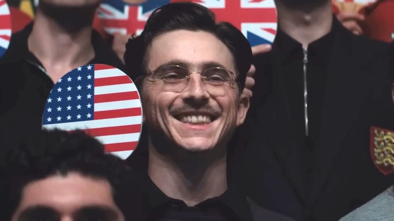 Timothée Chalamet as Marty Mouser holding up a table tennis paddle with an American flag emblem and smiling in Marty Supreme