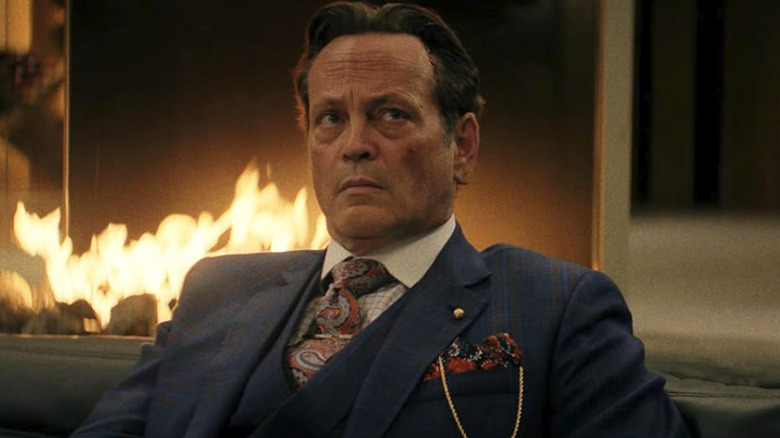 Nick wears a suit and sits in front of a fireplace in Mike & Nick & Nick & Alice