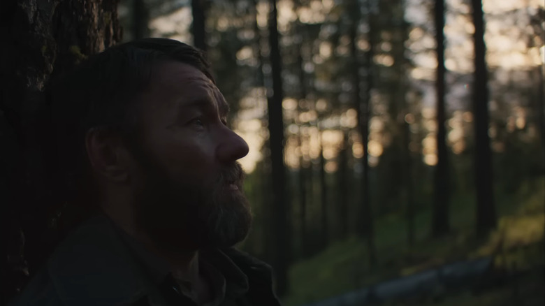 Joel Edgerton's Robert Grainier sits against a tree in a forest in Train Dreams