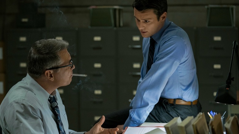 Holt McCallany's Bill Tench speaks to Jonathan Groff's Holden Ford, who sits on his desk in a room full of filing cabinets in Mindhunter