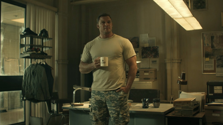Alan Ritchson's Jack Reacher stands in an office in his Army fatigues holding a mug in Reacher