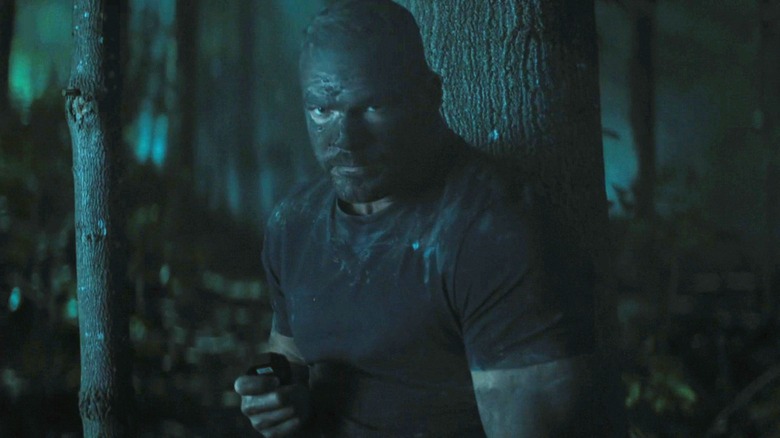 Alan Ritchson's Jack Reacher stands in a forest at night covered in mud in Reacher